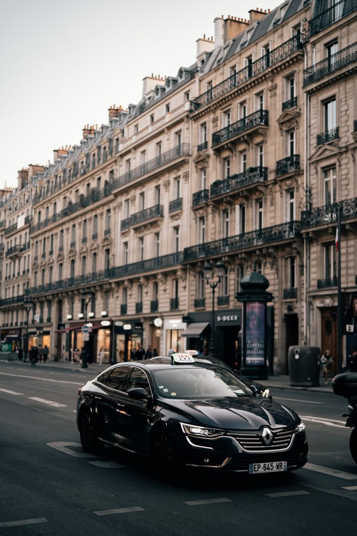 gallery-4 Taxi driving past historic buildings on a Parisian street, capturing urban charm and architecture.