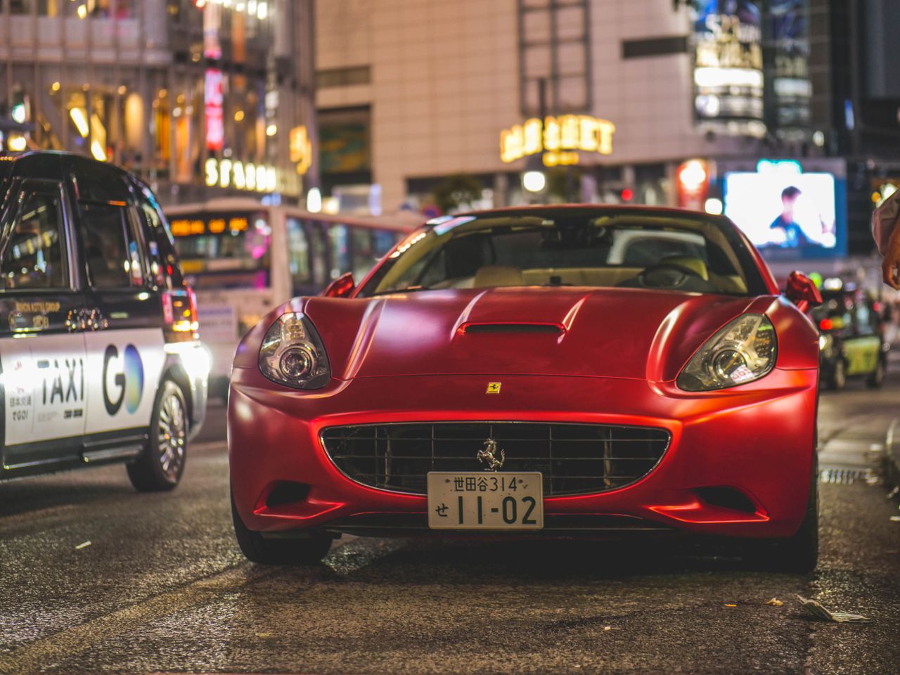 about-01 A sleek red Ferrari California parked on a busy city street at night, reflecting urban nightlife.