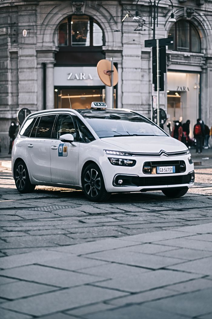 gallery-5 A modern white taxi car driving on a stylish city street with architectural backdrop.